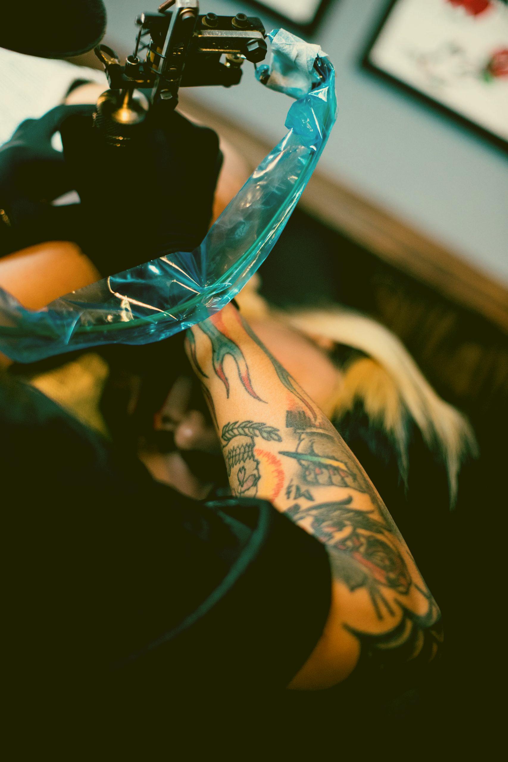 Imagen del brazo de un tatuador haciendo un trabajo. Se ve el cable de la máquina debidamente protegido con una lámina film por cuestiones higiénicas y sanitarias